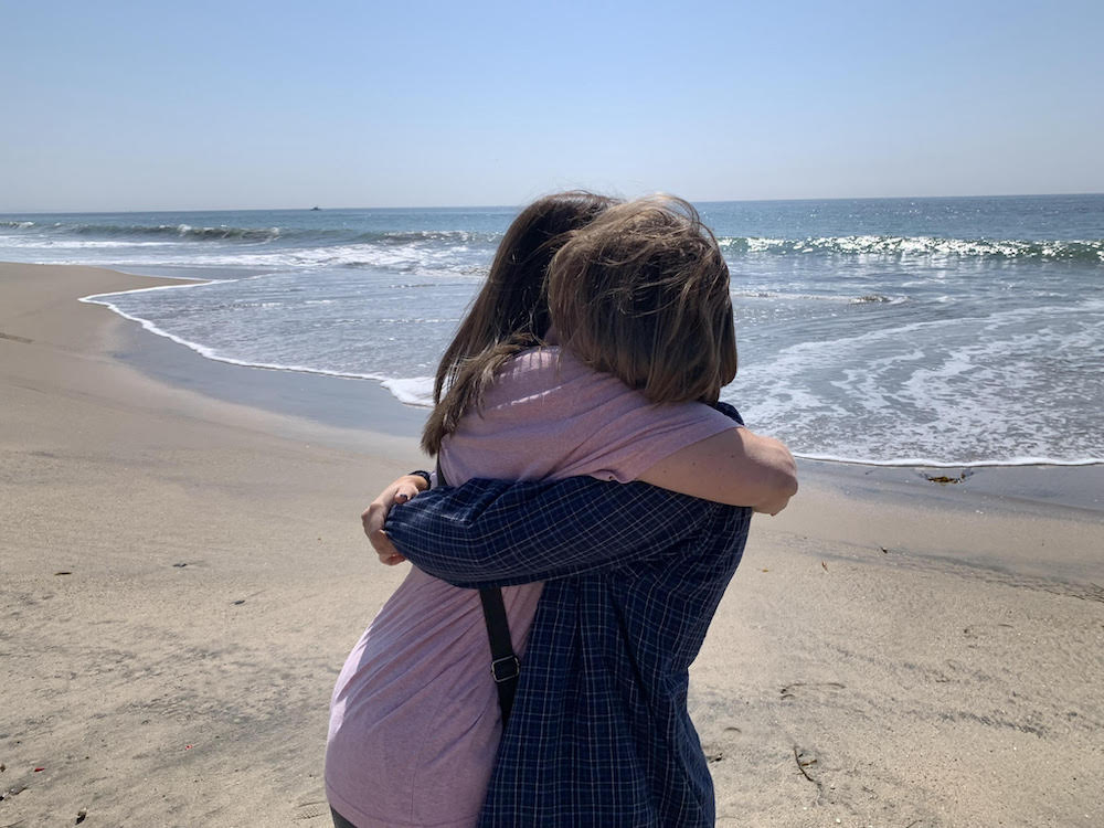 Two women embrace on the beach. One wears a pink T-shirt and one wears a long-sleeved plaid shirt.