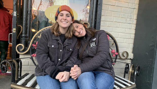 Two white women with sandy hair, black jackets, and jeans, lean into each other on a bench and smile