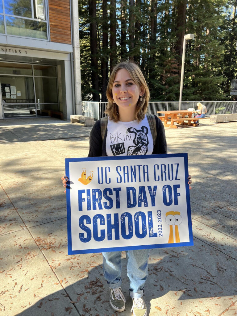 A young woman with light skin and shoulder-length blonde hair holds a sign that says "UC Santa Cruz First Day of School!" A campus building and trees are visible in the background.