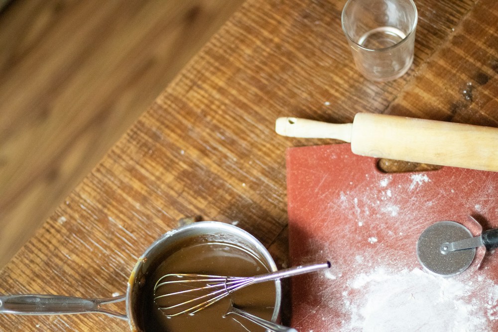 Remnants of baking on a countertop: a floured cutting board, rolling pin, cup, and small saucepan with brown sauce and a whisk in it