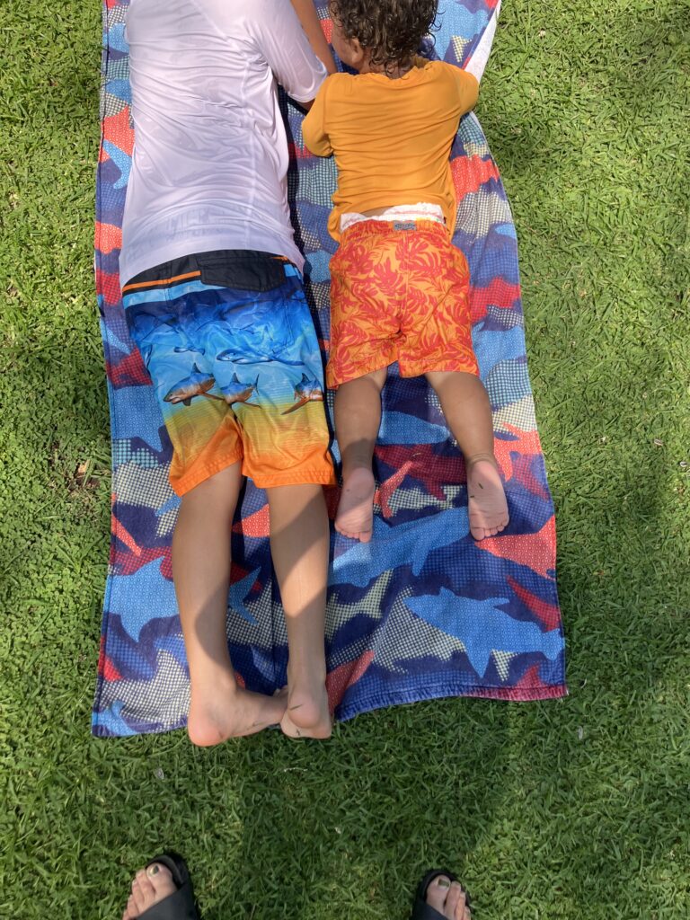 Two boys, ages 9 and 2, lay tummies-down on a towel on the grass