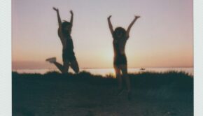 Two teen girls throw their arms up on a beach near sunset; they are seen in silhouette